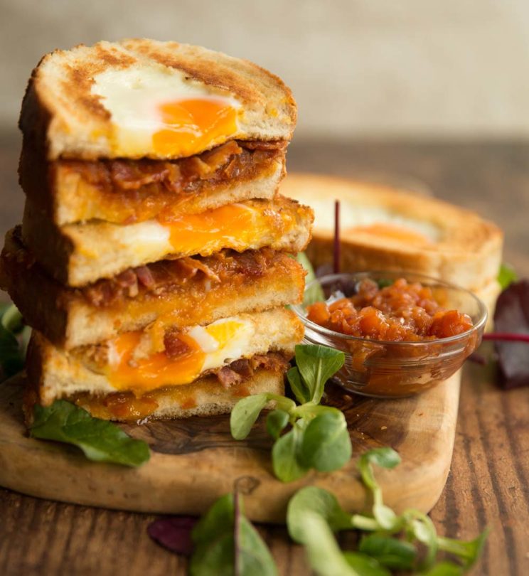 egg and bacon sandwiches stacked on top of each other on wooden board with salad to garnish