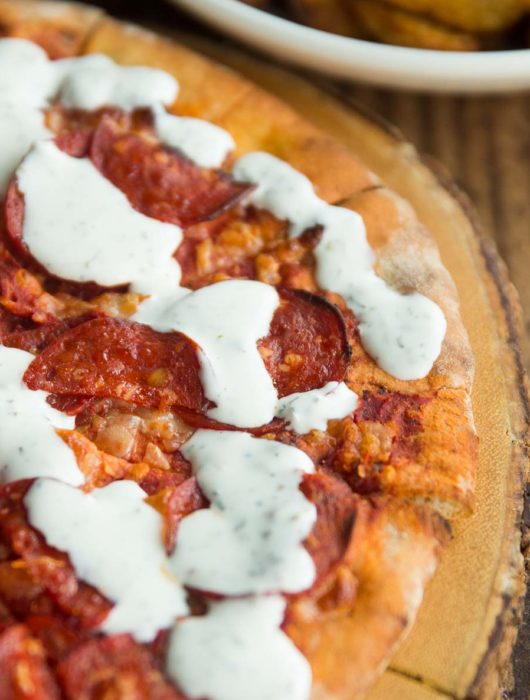 garlic and herb dip drizzled over pepperoni pizza on wooden board with potato wedges blurred in background