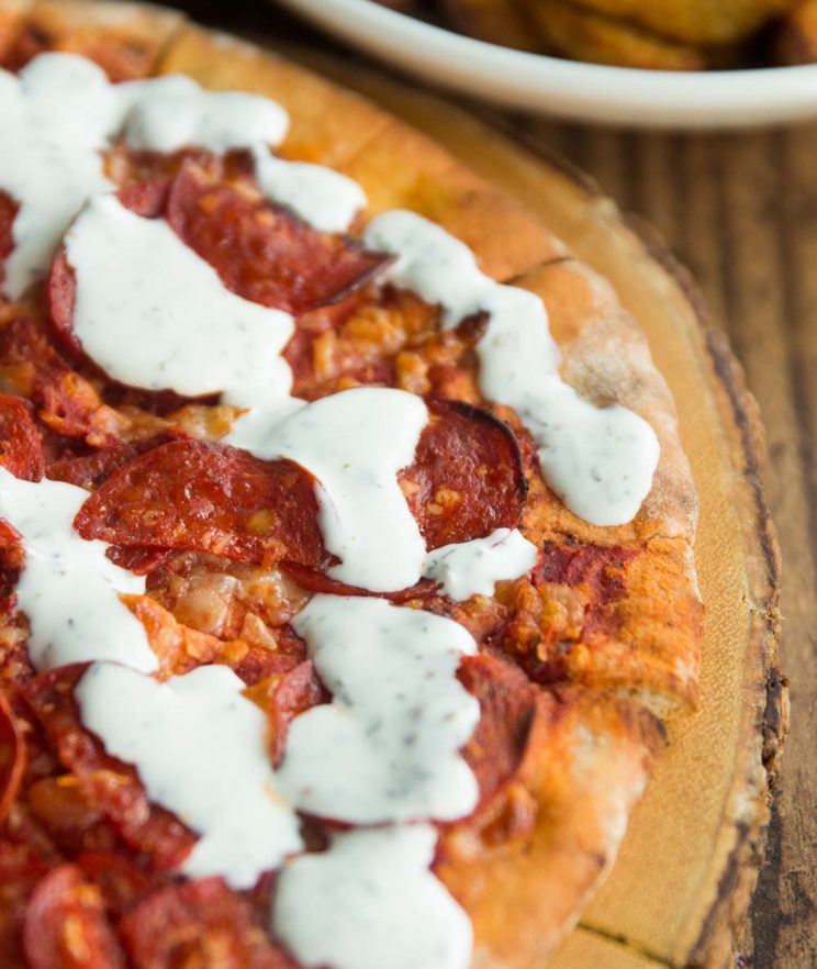 garlic and herb dip drizzled over pepperoni pizza on wooden board with potato wedges blurred in background