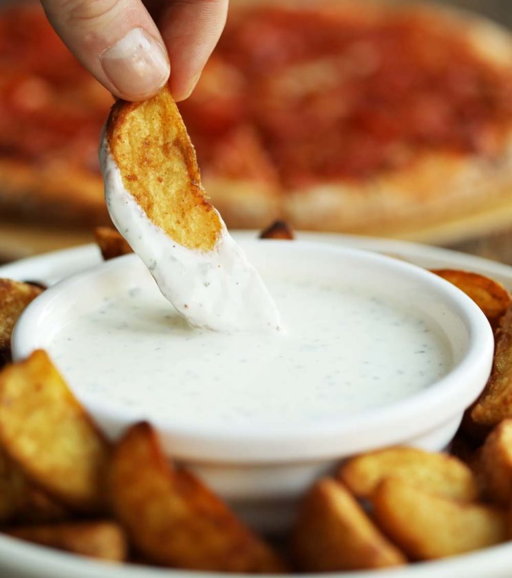 dunking potato wedge in garlic and herb dip with pizza blurred in the background