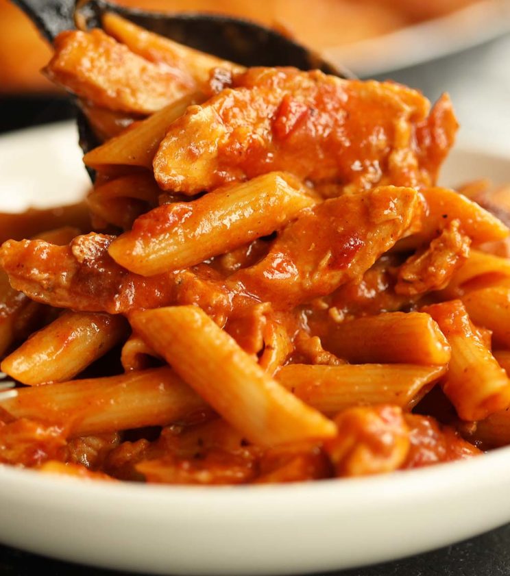 spoon pouring pasta into white bowl with skillet blurred in background