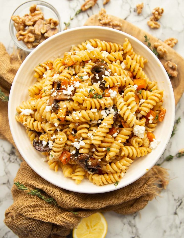 overhead shot of bowl of pasta on chopping board surrounded by walnuts and garnished with goats cheese