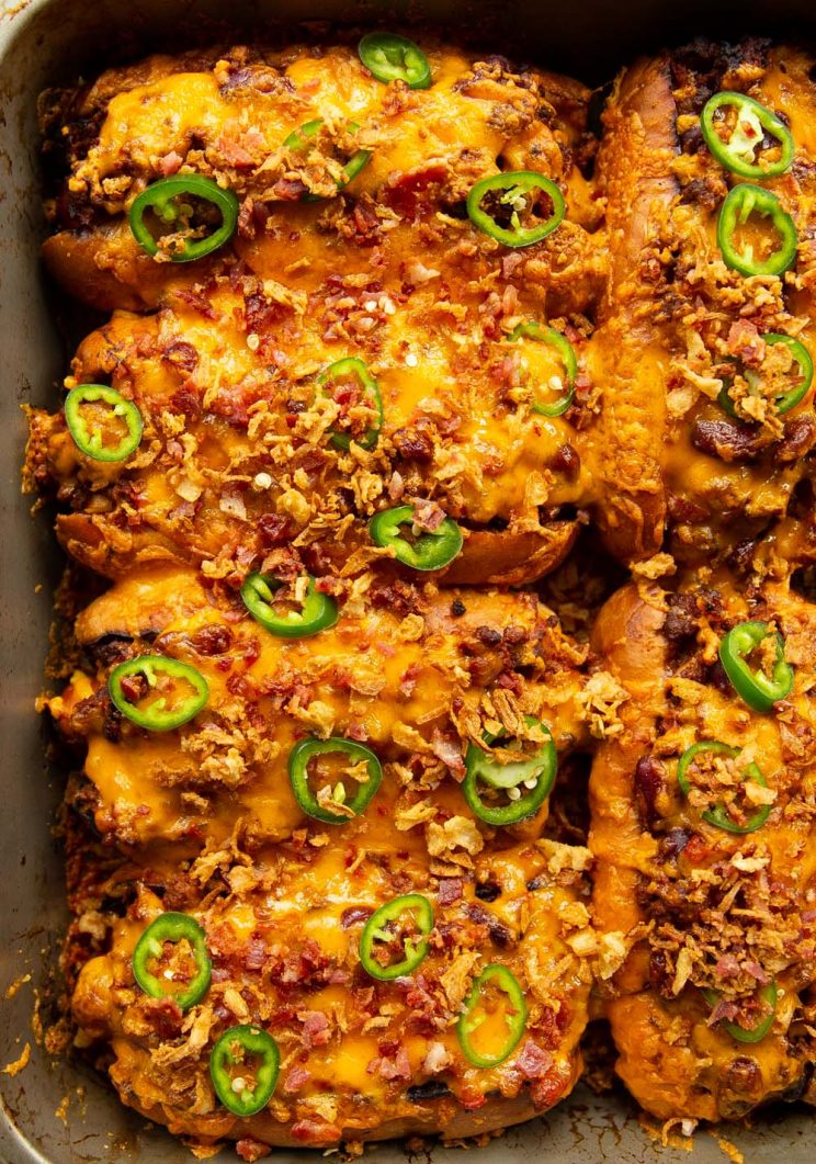 overhead shot of chili dogs garnished with jalapenos and crispy onion and bacon in baking tray
