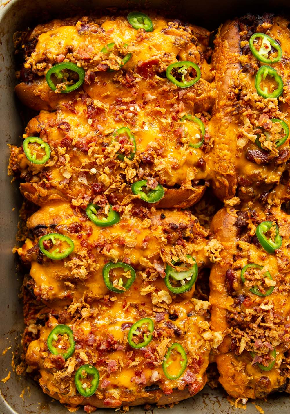 overhead shot of chili dogs garnished with jalapenos and crispy onion and bacon in baking tray
