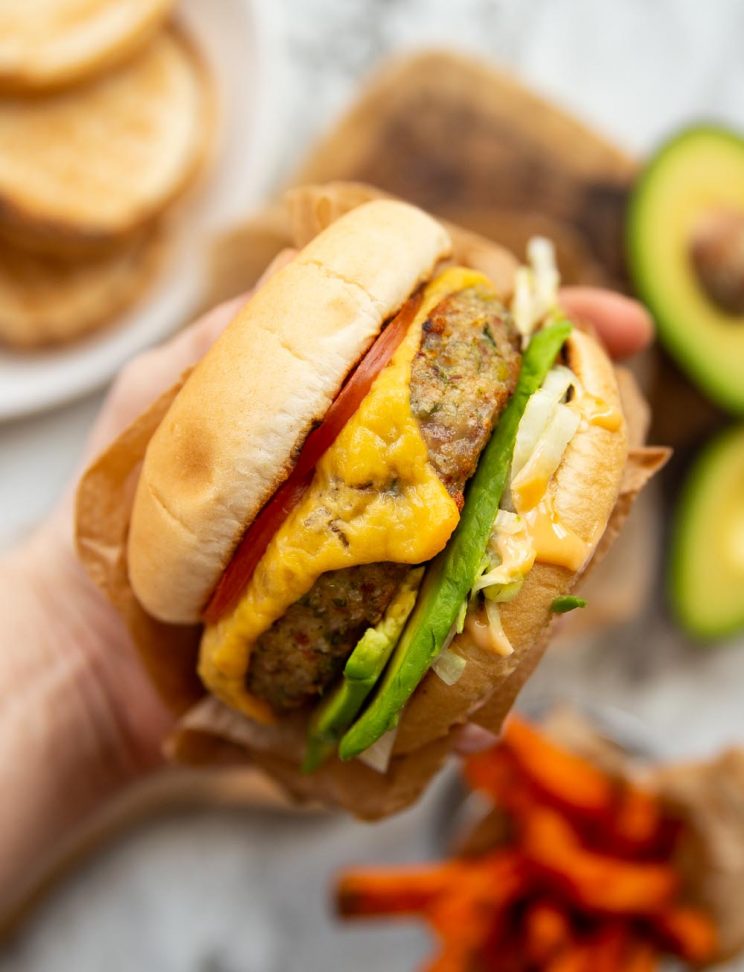 holding burger above chipping board with buns, avocado and sweet potato fries blurred behind