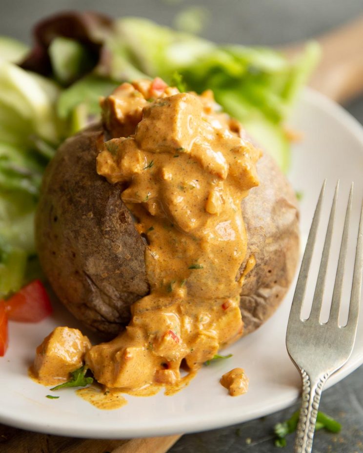 closeup shot of baked potato with chicken tikka on top with salad blurred behind