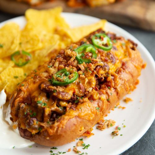 chili dog on white plate served with tortilla chips and more chili dogs blurred in background