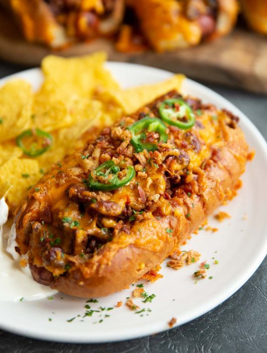 chili dog on white plate served with tortilla chips and more chili dogs blurred in background