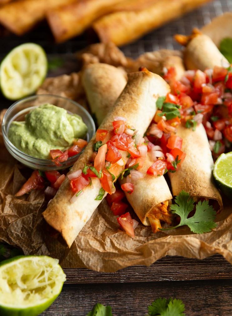 taquitos on parchment paper topped with salsa surrounded by limes and dip