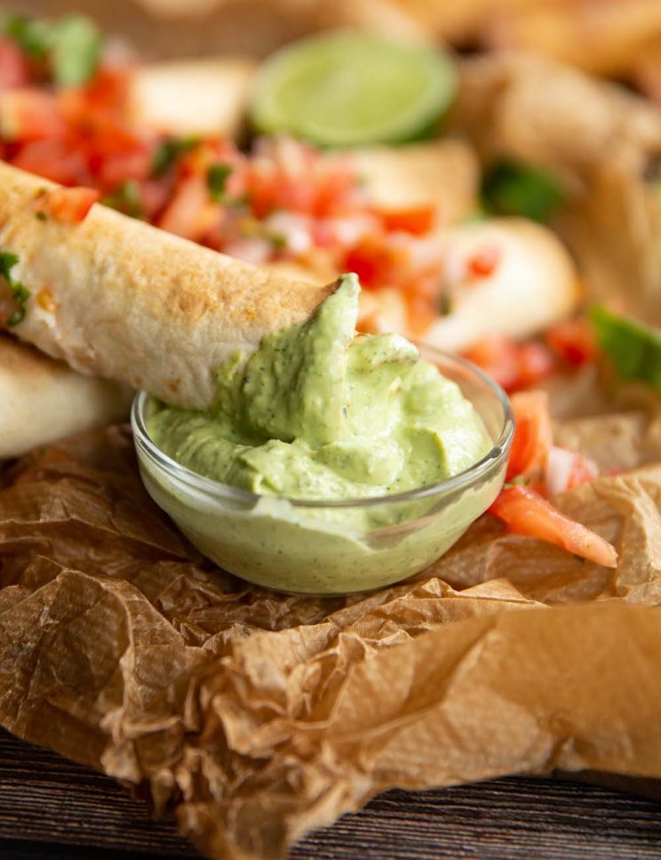 closeup shot of taquito dipped into glass pot of avocado dip