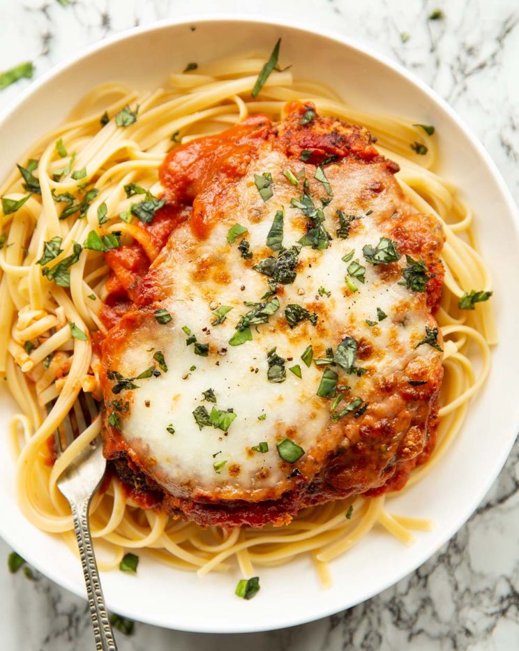 overhead shot of chicken parmesan served on spaghetti in white dish with silver fork digging in