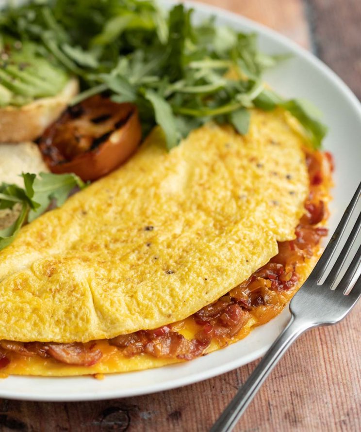 closeup shot of omelette on white plate with silver fork resting on it with salad blurred in background
