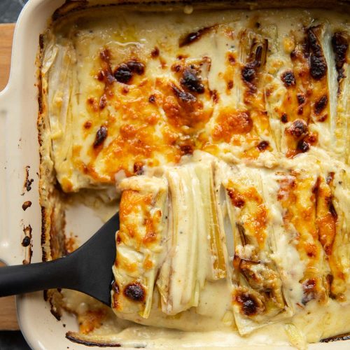 overhead shot of black plastic spatula digging into cheesy leeks in baking dish