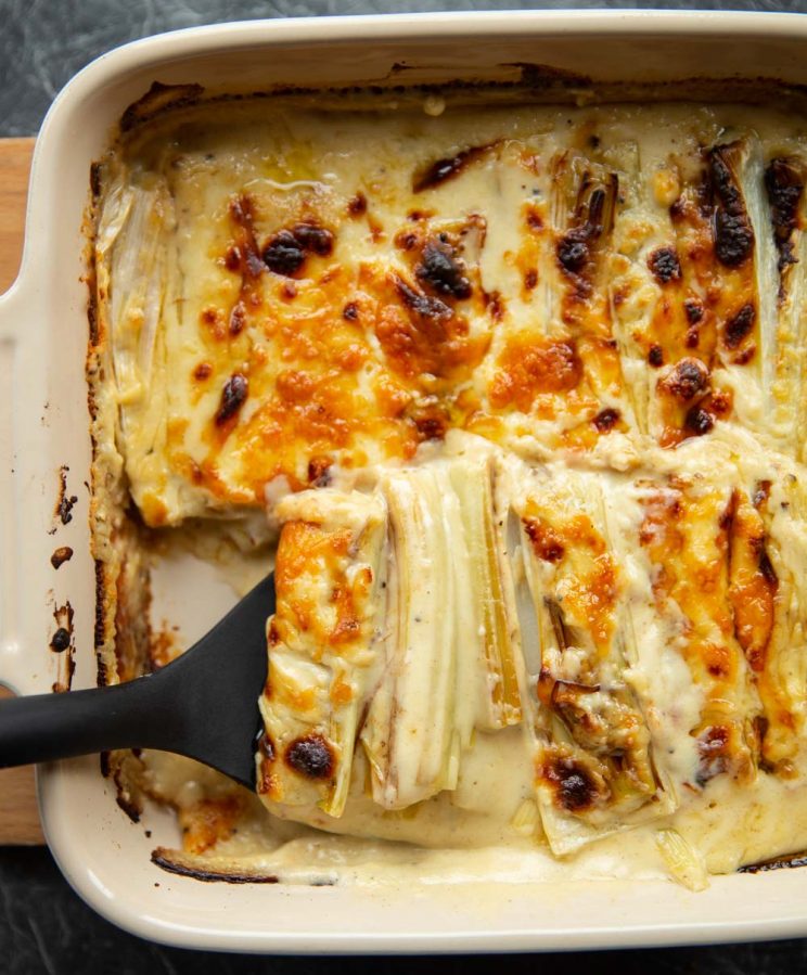 overhead shot of black plastic spatula digging into cheesy leeks in baking dish