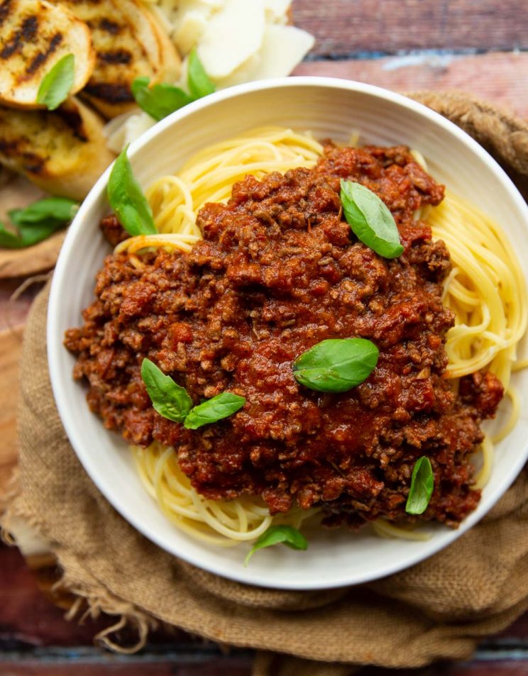 overhead shot of bolognese on spaghetti in white bowl with garlic bread blurred in corner