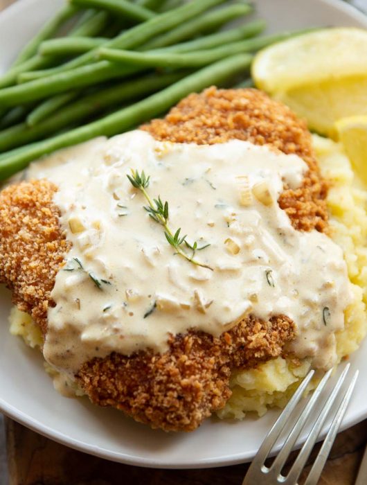 sauce drizzled over schnitzel on mash potato with green beans and lemon wedges blurred in background