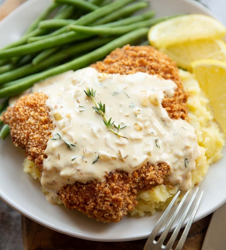 sauce drizzled over schnitzel on mash potato with green beans and lemon wedges blurred in background