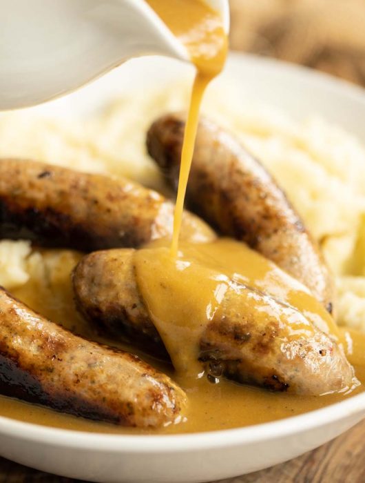 pouring gravy over sausages on mashed potato in white bow