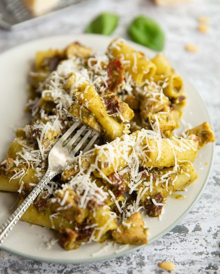silver fork digging into pasta on white plate with basil and pine nuts blurred in background