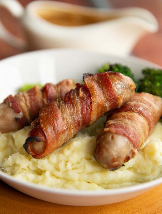 sausages on bed of mashed potato with jug of gravy blurred in background