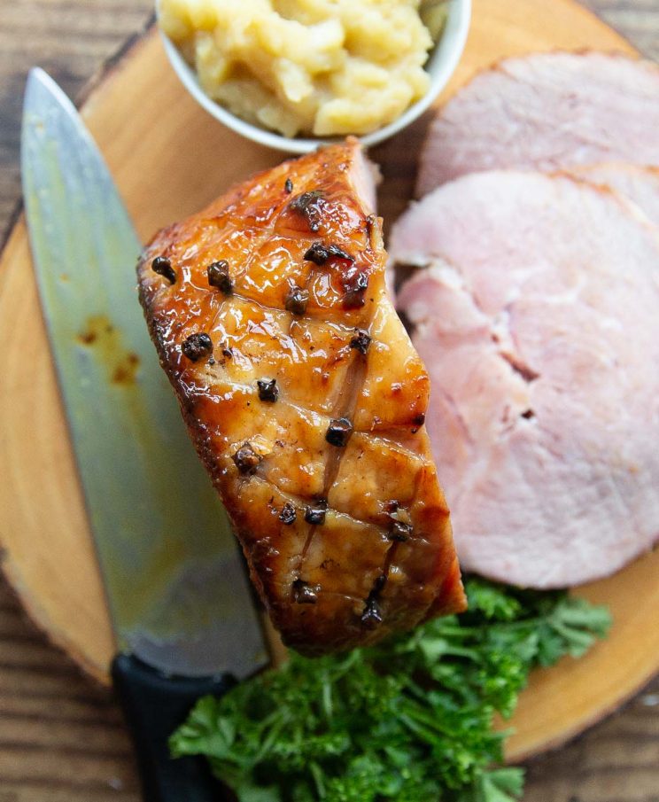 overhead shot of sliced ham on wooden board with knife and pot of apple sauce