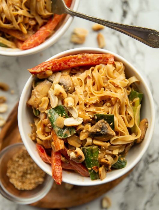 overhead shot of noodles in a white bowl surrounded by peanuts and sesame seeds