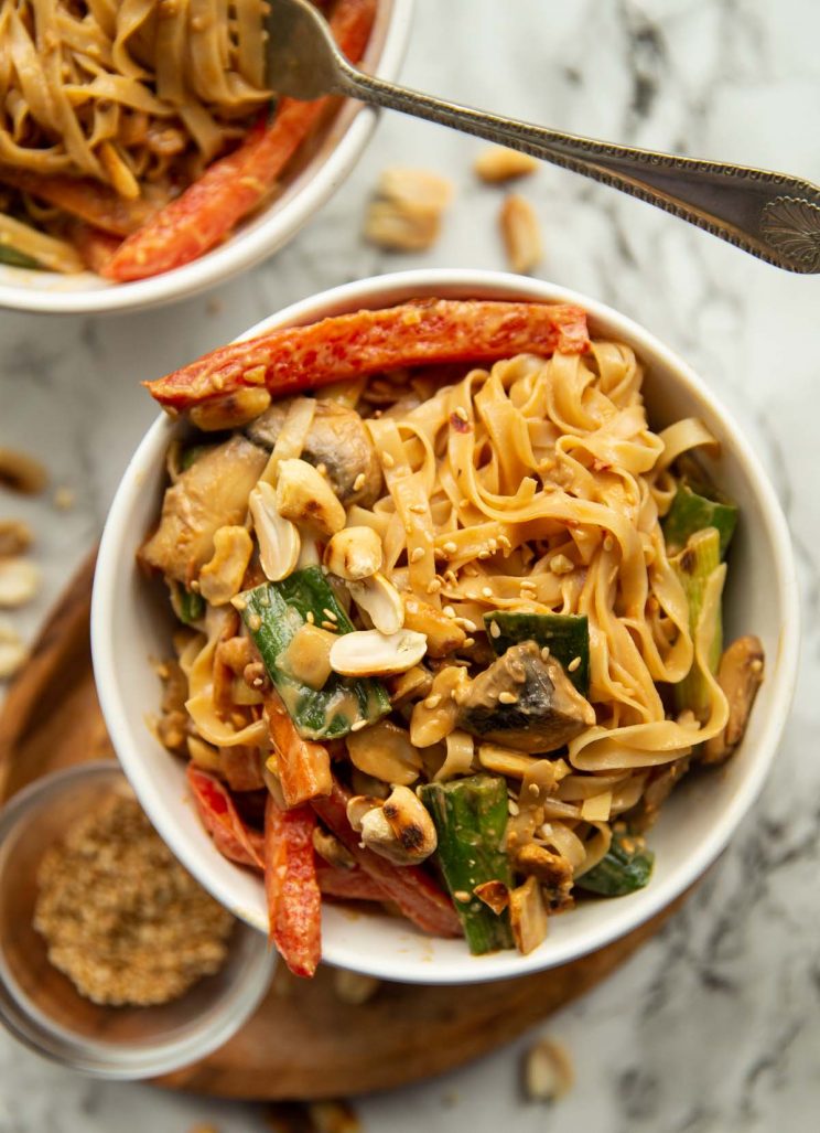 overhead shot of noodles in a white bowl surrounded by peanuts and sesame seeds