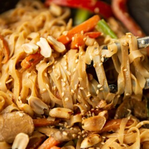 tongs twizzling noodles in wok garnished with peanuts and sesame seeds