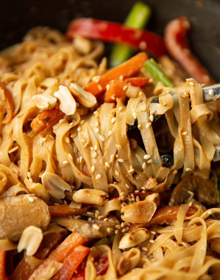 tongs twizzling noodles in wok garnished with peanuts and sesame seeds