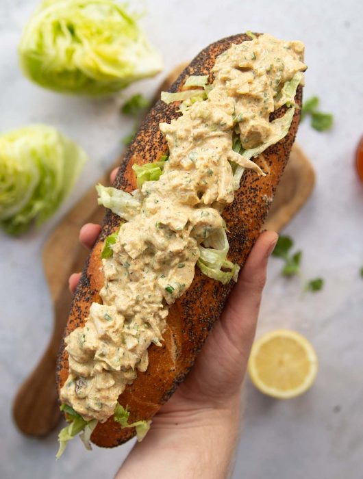 overhead shot of hand holding baguette filled with coronation chicken above garnish
