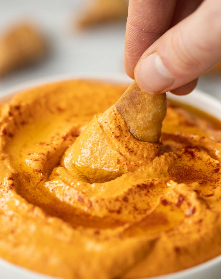 dunking a pita chip into small bowl of dip with more pita chips blurred in background