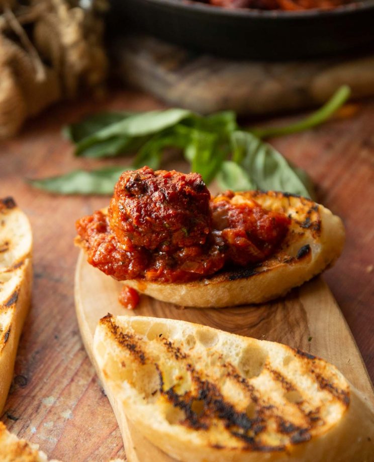 meatball on slice of garlic bread on chopping board with basil in background