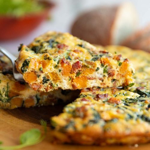 lifting slice of frittata above wooden board with salad and bread blurred in background