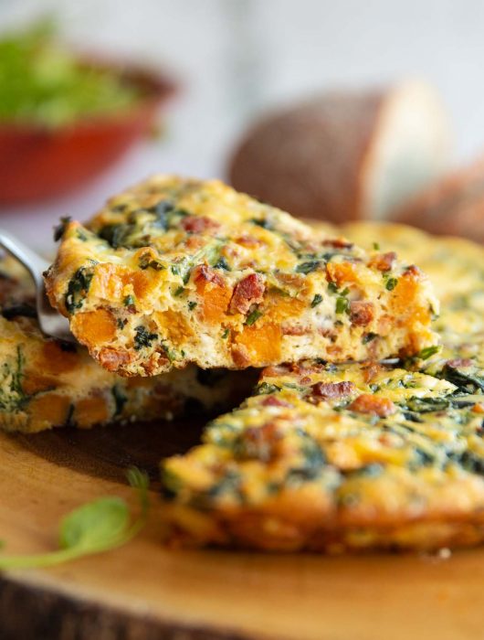 lifting slice of frittata above wooden board with salad and bread blurred in background