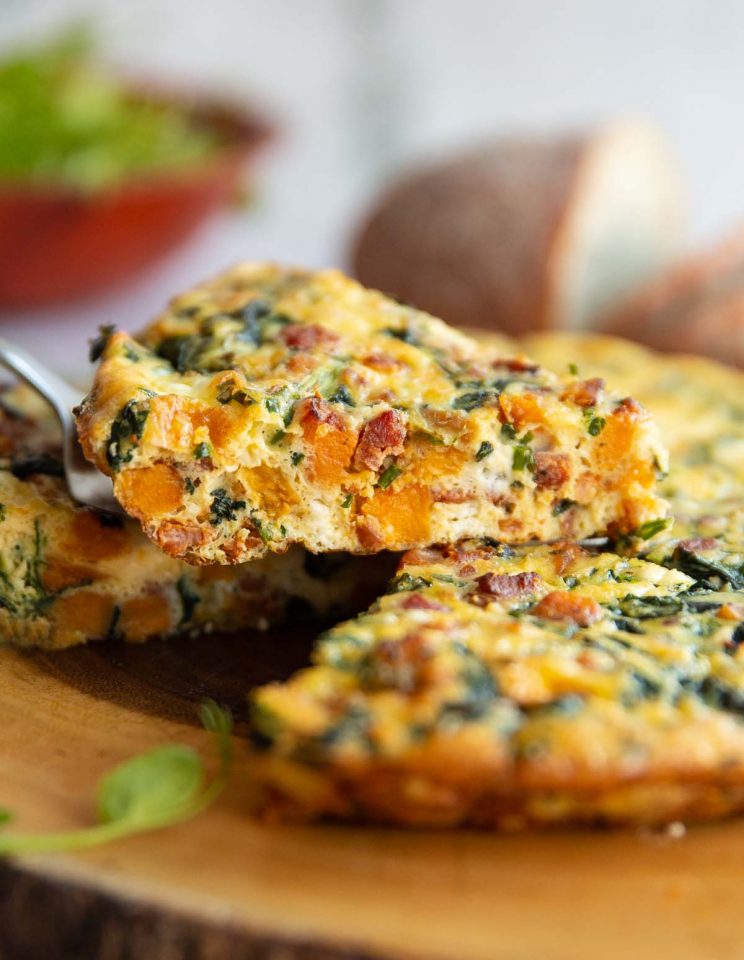 lifting slice of frittata above wooden board with salad and bread blurred in background