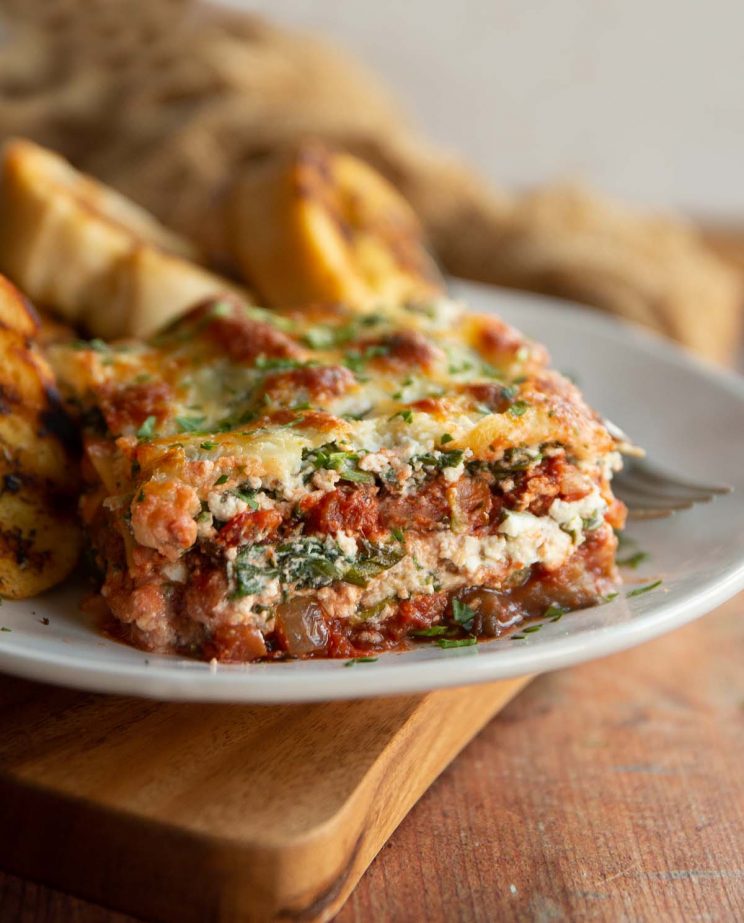 lasagne served on white plate with garlic bread blurred in background