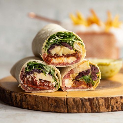 3 wraps stacked on each other on wooden board with dip and fries blurred in background
