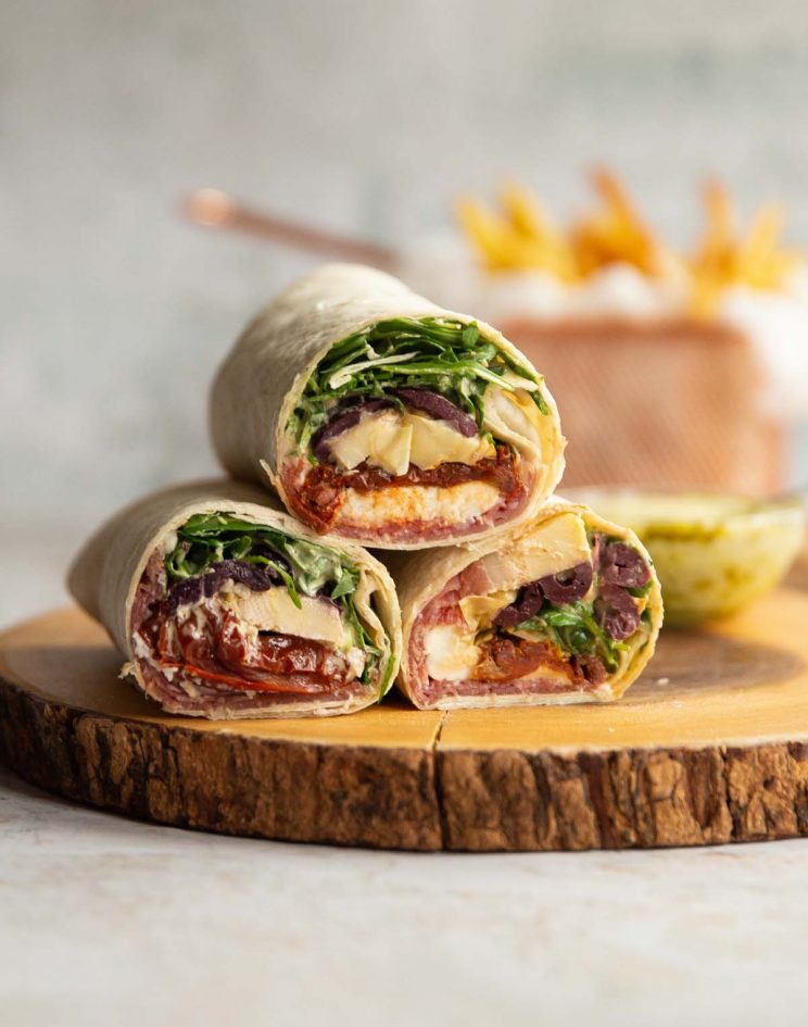 3 wraps stacked on each other on wooden board with dip and fries blurred in background
