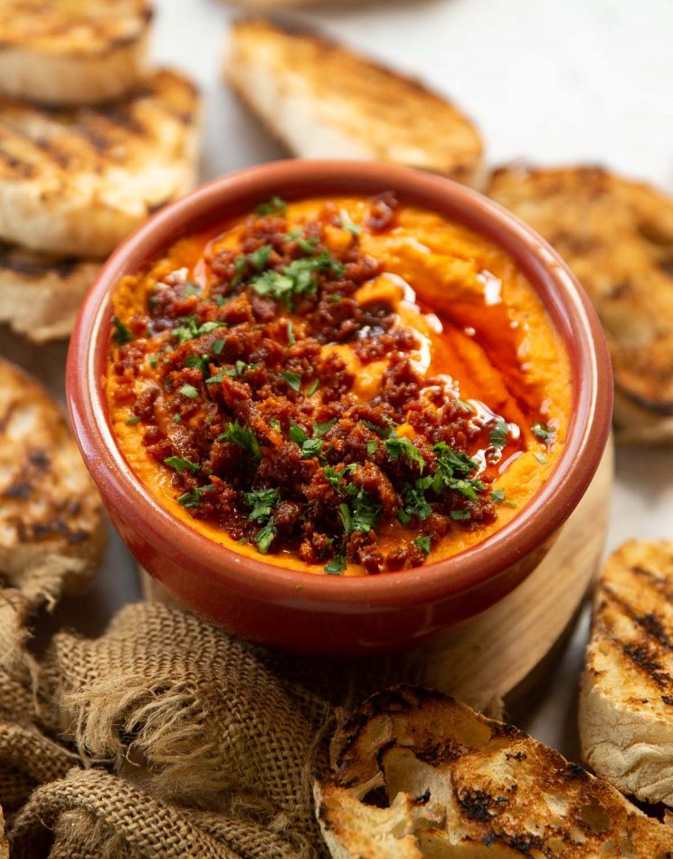 side shot of harissa dip in small red bowl surrounded by toasted ciabatta
