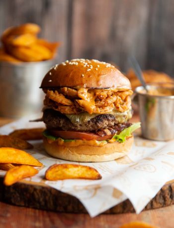 cowboy burger on shees of paper on wooden board surrounded by potato wedges