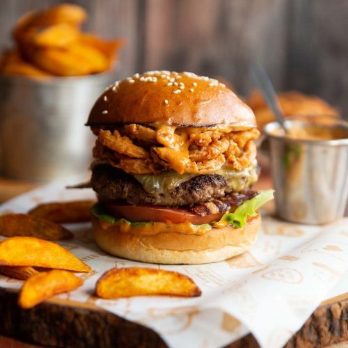 cowboy burger on shees of paper on wooden board surrounded by potato wedges