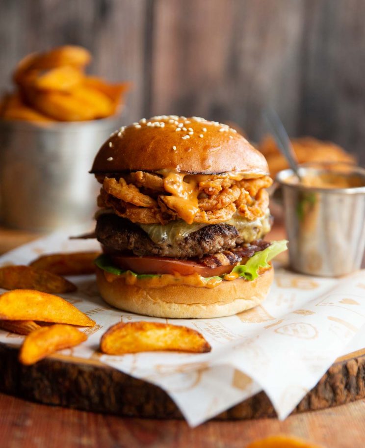 cowboy burger on shees of paper on wooden board surrounded by potato wedges