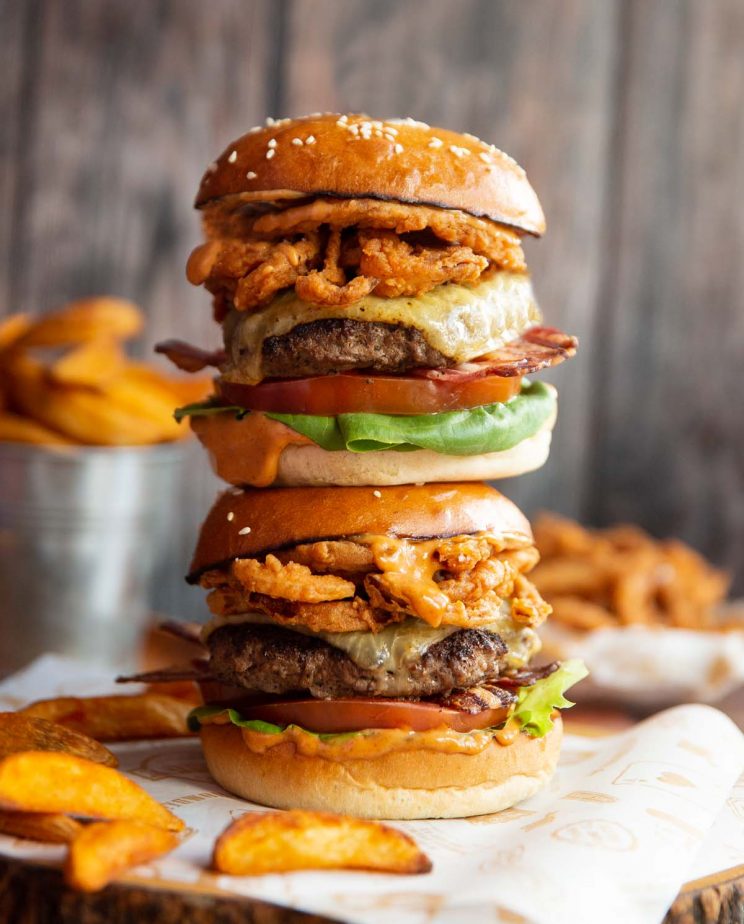 2 burgers stacked on top of each other on wooden board with fried onions and wedges blurred in background