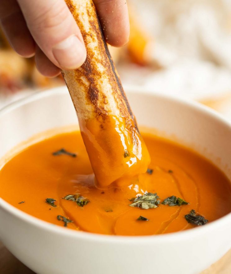 hand dunking grilled cheese roll up into small white bowl of tomato soup
