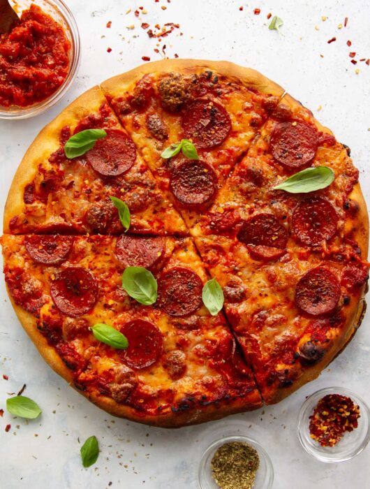 overhead shot of pizza sliced up on white background surrounded by chilli flakes and oregano