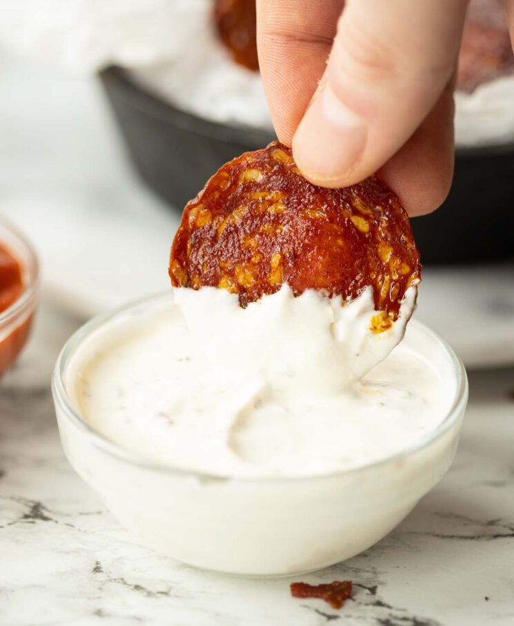 hand dunking pepperoni chip into small glass pot of dip