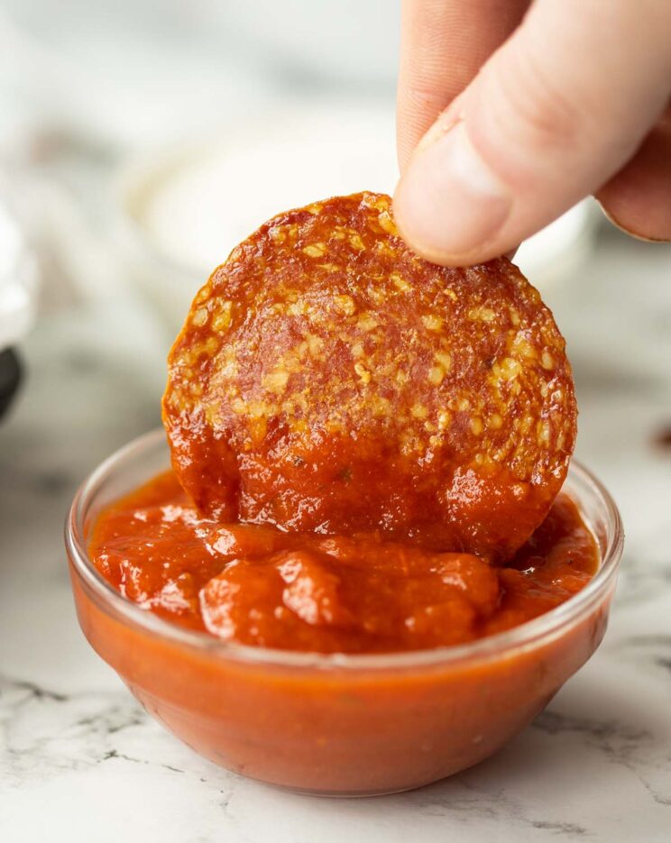 hand dipping salami chip into small pot of pizza sauce