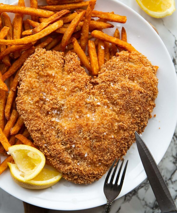 overhead shot of heart shaped chicken on white plate with fries and lemon wedges
