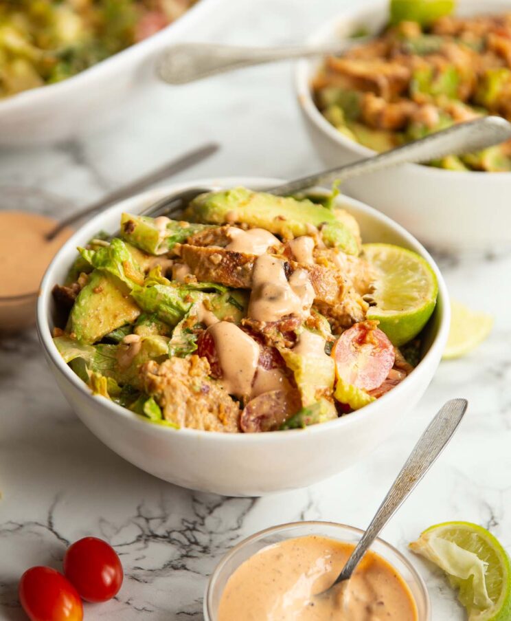 grilled chicken salad served in small white bowl with silver fork surrounded by garnish