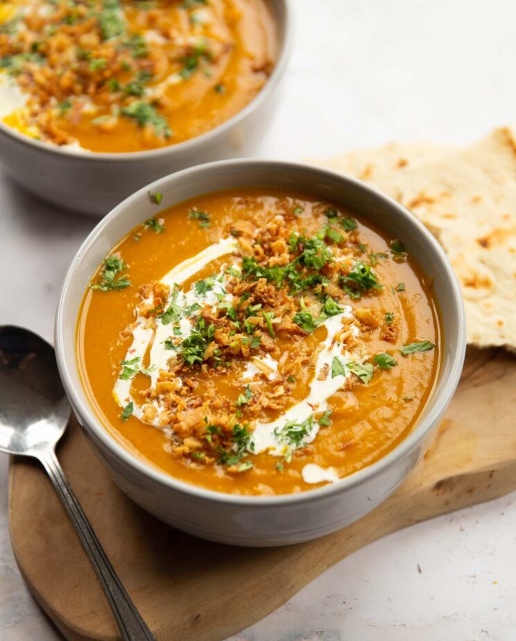 curried sweet potato soup served in large grey bowl on wooden board with silver spoon and flatbread